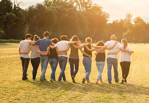 Group of friends with their arms around each other and walking across a meadow towards the sun